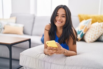 Obraz na płótnie Canvas Young hispanic woman using smartphone lying on sofa at home