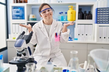 Fototapeta premium Hispanic girl with down syndrome working at scientist laboratory clueless and confused expression with arms and hands raised. doubt concept.