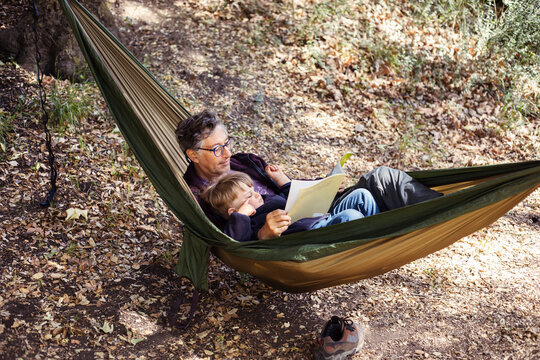 Grandfather reading book for grandson lying on hammock in forest