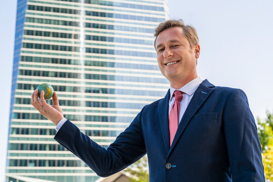 Smiling Businessman Holding Small Globe Ball In Front Of Office Building