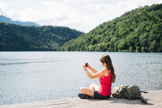 Woman photographing Lake Levico through smart phone sitting on jetty