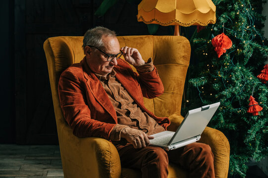 Senior man using laptop sitting by Christmas tree at home - Powered by Adobe
