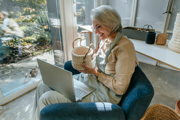 Happy mature craftswoman showing crochet basket on video call through laptop in workshop