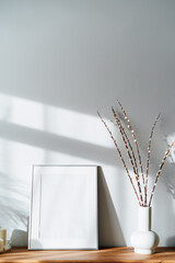 Modern minimalist style interior with white poster mockup, candles and blooming branches of the pussy willow in vase on wooden console under sunlight and shadows on a white gray wall. Selective focus.