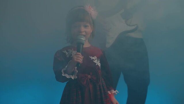 Little girl on stage in vintage dress, she sings into microphone and dances, her father plays an electric guitar. Color music is shining and smoke is billowing. Family time, creativity and hobbies