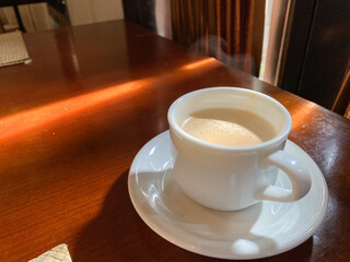 close up of coffee cup on table at direct sunlight. morning coffee with steam in white cup