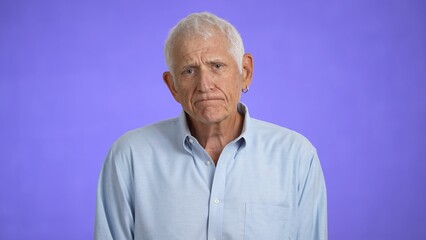 Sad upset worried disturb displeased shocked elderly gray-haired man 70s wears blue shirt sigh surprise put hands on face isolated on solid purple background studio portrait