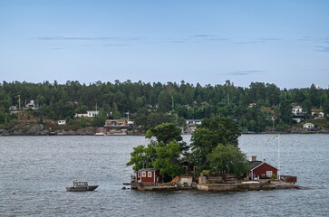 Fototapeta na wymiar Sweden, Stockholm - July 16, 2022: Morning approach by ship from Baltic. Very small Kattholmen Island with a couple buildings and green foliage under blue sky. Small boat.