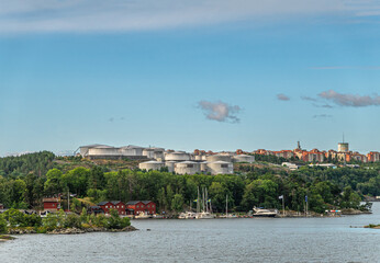 Sweden, Stockholm - July 16, 2022: Shoreline under Bergs oljehamn, oil port, in Nacka. Many oil tanks under blue morning sky. Historic red buildings and ships in small yacht port