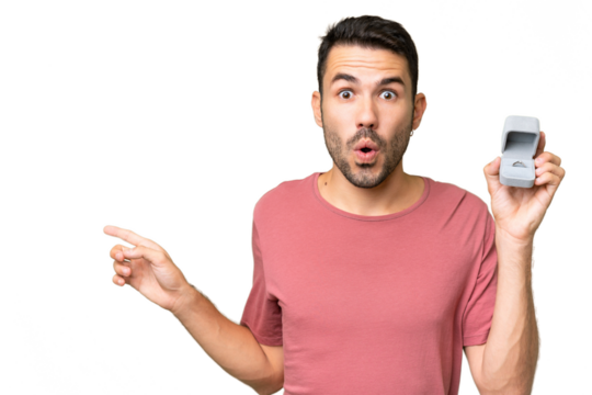 Young handsome caucasian man holding a engagement ring over isolated background surprised and pointing side