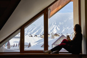 Woman being cold sits on windowsill, warming up with tea mug, looking at snow covered mountains....