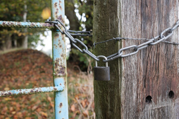 Padlock and chain linking an old metal gate and a wooden pole with barbed wire, blocking the passage to a private area. 