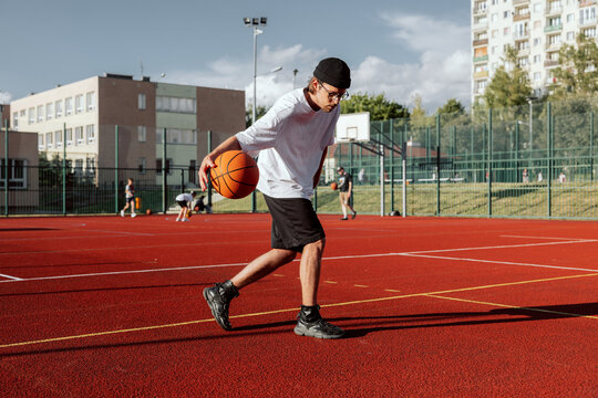hippster guy in sportswear and glasses showing skills at basketball court trying to make slam dunk during game. Man having fun with guys company spending free time outside.