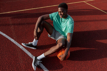 African american baketball player wearing sportswear sitting on basketball court holding basketball ball in hands resting relaxing having break during game with friends company.