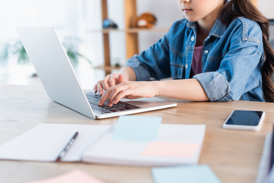 cropped view of girl typing on laptop near blurred copybook while studying at home.