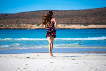 Fototapeta na wymiar beautiful long-haired girl in a dress soaks her legs in turquoise water on a paradise beach at turquoise bay in cape range national park near exmouth, western australia; beach with red cliffs in the b
