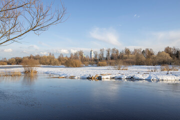 Naklejka na ściany i meble March sunny day by the river. A picturesque landscape, early spring, a river with snow-covered banks, dry grass and bushes. Church in the background. The first thaws, the snow is melting
