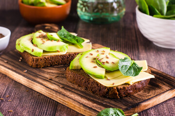 Sandwiches on rye bread with cheese, avocado, spinach and flax seeds on a wooden board on the table.
