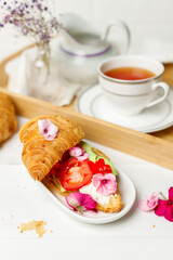 Breakfast. Croissant with cream cheese and edible flowers. Wooden tray with a cup of tea, creamer. Close-up, vertical
