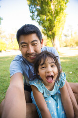 Asian dad taking selfie with his beautiful daughter. Handsome man spending vacation with his little girl in park both sitting on grass laughing looking at camera. Leisure, active rest outdoors concept