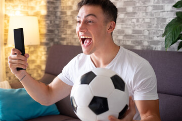 Excited young boy soccer fan watching a game on television holding a soccer ball in hand and a...