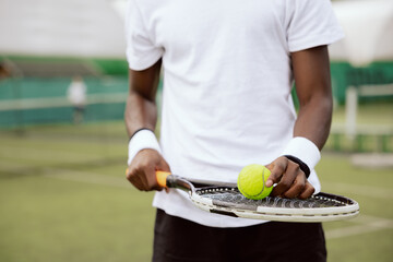Close-up of the of African-looking man holding tennis racket and ball. Tennis player is wearing white T-shirt and black shorts and has wristbands on hands. He is playing tennis and is ready to serve.