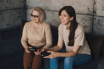 Cheerful adult woman playing video games with her mother while sitting on the sofa. Family playing video game. Having fun on weekend. Happy family, childhood.