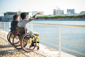 Side view of family in wheelchairs spending time near water. African American man and Caucasian woman in casual clothes, looking at water. Love, affection, happiness concept