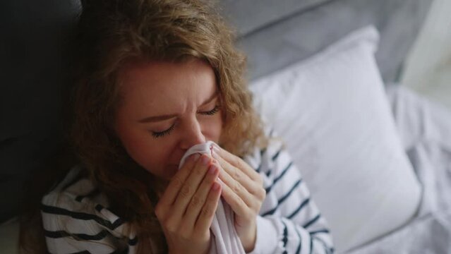 Woman sick with flu coughing and blowing her nose on her bed at home having sore throat, fever and headache. Young caucasian female suffering from coronavirus Covid19 while isolating touches forehead