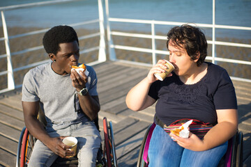 Cheerful biracial couple drinking coffee on sunny day. African American man and Caucasian woman in wheelchairs on embankment, drinking hot beverage from cups. Snack, relationship, happiness concept