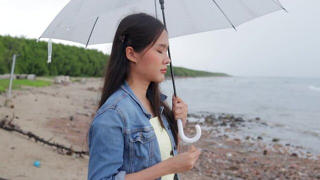 Beautiful Asian woman holding a white umbrella standing at the sea beach She sadly threw her wedding ring into the sea. Sad woman alone at the beach during the rain.