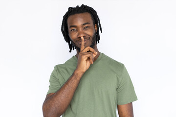 Portrait of pleased African American man shushing. Secretive young male model with braided dark hair in green T-shirt looking at camera with finger on lips, asking to be quiet. Secret, silence concept