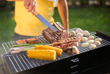 Close-up of man grilling tasty meat and vegetables on sunny day. Man in yellow T-shirt turning roasted ribs on BBQ grid. BBQ, cooking, food concept