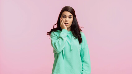 dissatisfied and brunette woman in turquoise hoodie touching cheek isolated on pink