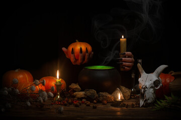 Halloween night. Witches hands with black long nails at work above a cast-iron pot among pumpkins,...