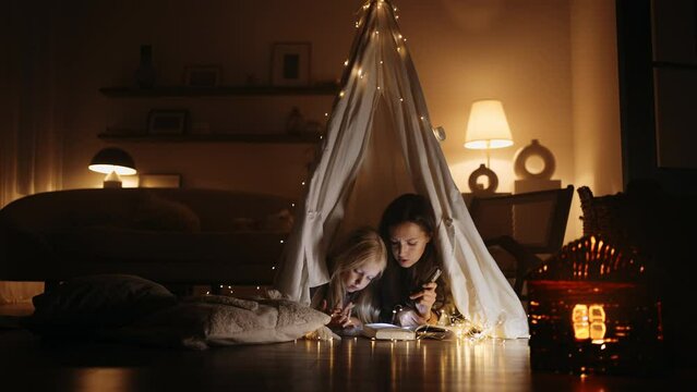 A young hispanic mother and daughter reads a book with a flashlight lying in a tent at home in the dark, magical decorations of the house. Cozy atmosphere for mother and daughter