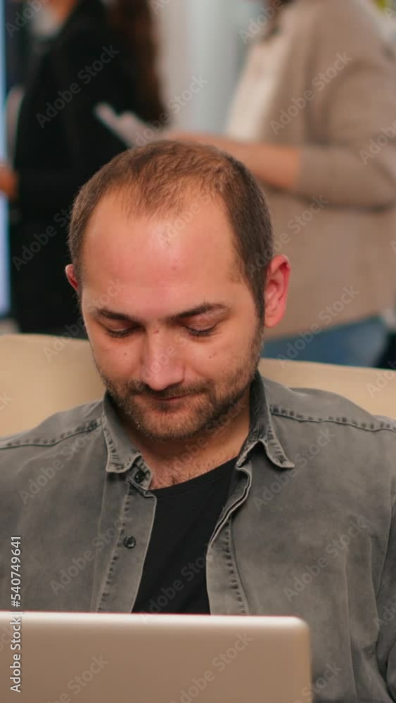 Poster manager sitting on couch in front of camera smiling while multiethnic team working in background wit