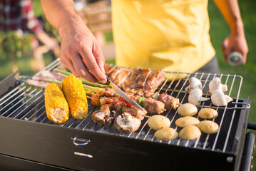 Close-up of man grilling meat and vegetables on sunny day. Man in yellow T-shirt turning products on BBQ grid. BBQ, cooking, food concept
