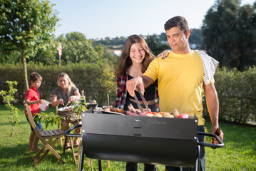 Content father grilling meat and vegetables with daughter. Man in yellow T-shirt standing near BBQ grid with smiling teenage girl. Wife and son in background. BBQ, cooking, food, family concept