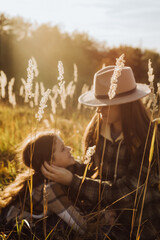 Selective focus of caring young mother and happy little daughter in hat sitting in old grass on background amazing sunset or sunrise. Connection with nature and environmental protection concept