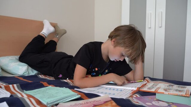 Preteen child doing homework exercise lying on bed at home. Using smartphone searching information. Mess in children room. School classes, education, handwriting test. Remote education. E-learning