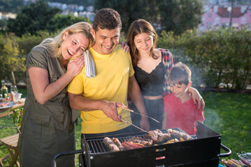 Loving father grilling meat and vegetables with family. Dark-haired man in yellow T-shirt standing near BBQ grid with little son, wife and teenage daughter. BBQ, cooking, food, family concept