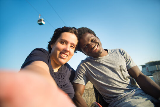 Smiling biracial couple taking selfie on embankment. African American man and Caucasian woman in wheelchairs on embankment, looking at camera. Love, relationship, social media concept