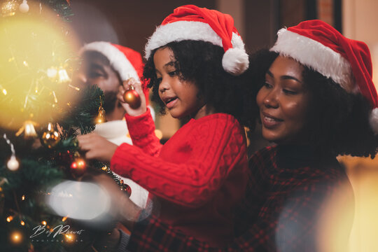 African American families happy and laughing celebrate Christmas eve sitting in leaving room decorations with a green Christmas tree and lighting, giving a surprise red gift box.