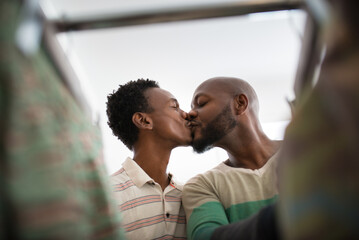 Close-up of African gay couple kissing at clothing store. Low angle view of two loving men standing near hangers with clothes choosing shirts enjoying time together. Same sex love and shopping concept