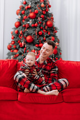 Obraz na płótnie Canvas young smiling father with baby boy are having fun on red sofa by the Christmas tree at home. Dad and son with the same pyjamas celebrating Xmas and New Years family holiday vacation. Fathers day
