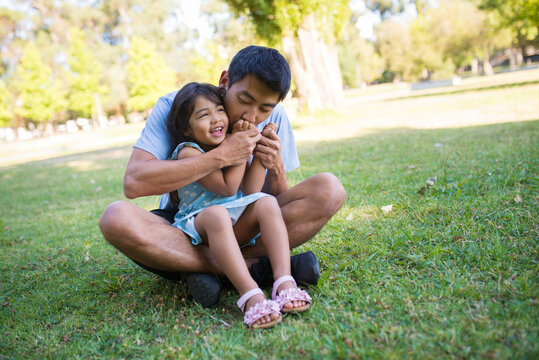 Portrait of happy Asian man holding little girl on his knees. Young dad sitting on green grass in public park hugging his beautiful daughter and kissing her hands. Parenting and summer rest concept