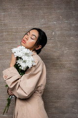 Fashionable asian woman in trench coat holding chrysanthemums and closing eyes on abstract brown background