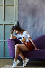 A beautiful stylish girl dressed in a skirt and T-shirt is sitting on a bright purple sofa. Loft interior.