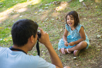 Portrait of little Asian girl and father taking photo of her. Beautiful girl in summer dress sitting on grass looking at fathers photo camera laughing. Parenthood and spending time together concept